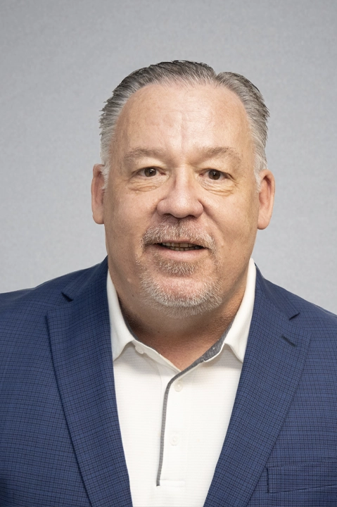 Portrait of a middle-aged man in a blue blazer and white collared shirt against a gray backdrop, looking at the camera with a neutral smile.